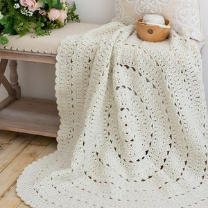 May include: An off-white, circular crochet blanket featuring an intricate, repeating openwork pattern with a scalloped edge. The textile is crafted from yarn and displays a textured, handmade design that radiates from the center outward in concentric circles.