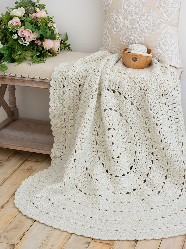 May include: An off-white, circular crochet blanket featuring an intricate, repeating openwork pattern with a scalloped edge. The textile is crafted from yarn and displays a textured, handmade design that radiates from the center outward in concentric circles.