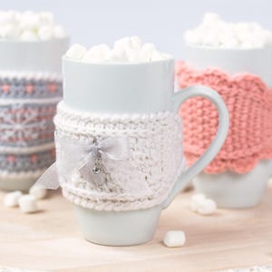 Puede incluir: Tres tazas de cerámica blanca llenas de chocolate caliente y malvaviscos, cada una con una funda de ganchillo única. La taza central tiene una funda blanca con un lazo de cinta gris y un dije de cristal, las otras tienen diseños de copos.