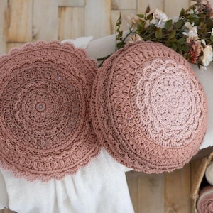 May include: Two round, dusty rose crochet cushions featuring intricate, concentric circular patterns with scalloped edges. The textured yarn work displays a detailed, repeating stitch design that radiates from the centre of each cushion.