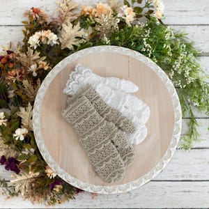 May include: Two pairs of handmade mittens, one beige and one white, rest on a wooden tray with a decorative lace border. The tray is surrounded by fall-themed floral arrangements with a rustic aesthetic.