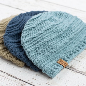May include: Three crocheted beanies in different shades of blue and brown. The beanies are stacked on top of each other. The top beanie has a brown leather tag with the letters "K.H." on it.