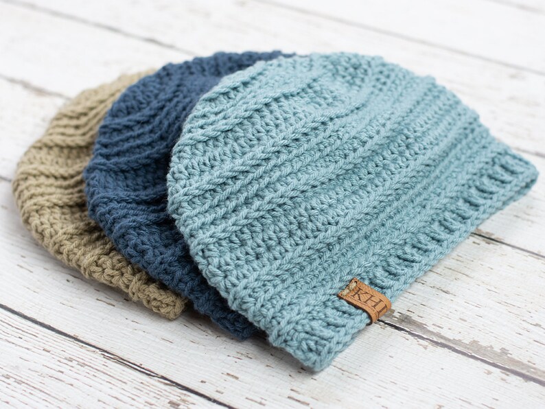 May include: Three crocheted beanies in different shades of blue and brown. The beanies are stacked on top of each other. The top beanie has a brown leather tag with the letters "K.H." on it.