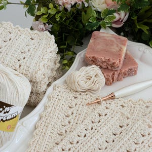 Crochet Primrose and Proper cream dishcloth with yarn, soap, crochet hook, flowers, and white tray on a wood surface.