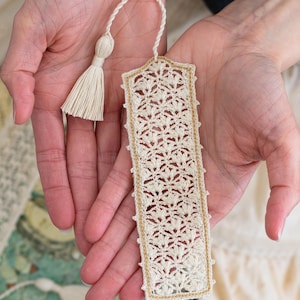 May include: Cream-colored, handmade crochet bookmark with a delicate floral pattern and a gold border. The bookmark has a matching tassel and is held in open hands. A beautiful, handcrafted accessory for book lovers.