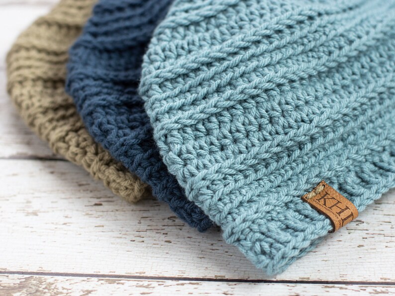 May include: Three crocheted beanies in different colors: green, blue, and light blue. The light blue beanie has a brown leather tag with the word "KHL" on it.