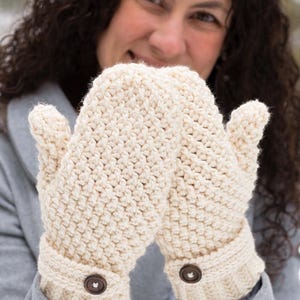 Könnte beinhalten: Cremefarbene Häkelhandschuhe mit einem strukturierten Stichmuster. Die Handschuhe haben gerippte Bündchen und sind mit braunen Knöpfen versehen. Das Bild zeigt eine Nahaufnahme der Handschuhe, die ihre handgefertigte Qualität und ihr gemütliches Design hervorhebt.