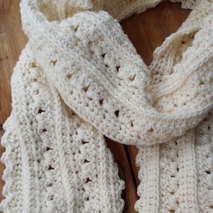 May include: A cream-colored crocheted scarf featuring a repeating vertical pattern of textured stitches and openwork details. The scarf has a scalloped edge along the length and is constructed from a soft yarn with a consistent off-white hue.