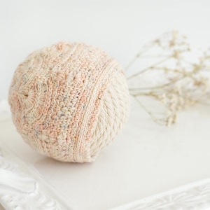 May include: A spherical ornament covered in a textured, hand-knitted fabric featuring a variegated peach, cream, and light blue speckled yarn pattern. The top section displays a circular knit design, while the lower half shows a ribbed texture.