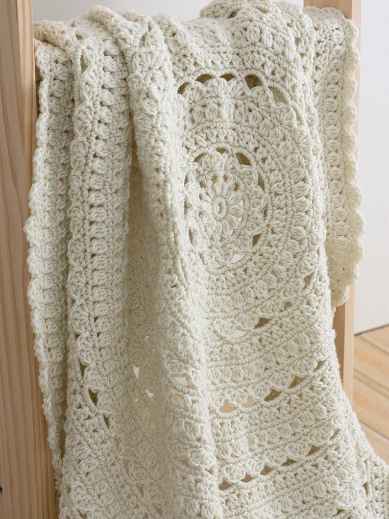 May include: A cream-colored crochet blanket featuring a central circular mandala pattern with intricate textured stitches, including puff stitches and openwork shells, finished with a scalloped border along the edges of the soft yarn fabric.