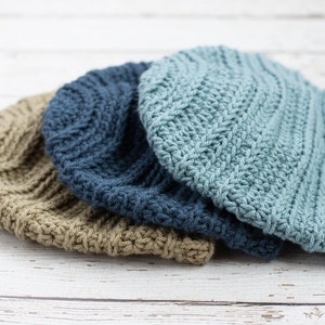May include: Three crocheted beanies in different colors: beige, blue, and light blue. The light blue beanie has a brown leather tag with the letters "KCH" on it.