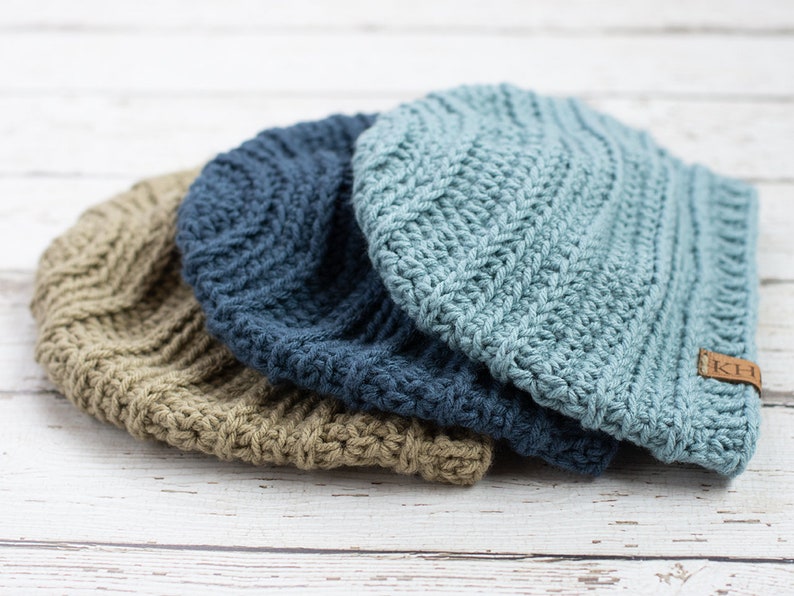 May include: Three crocheted beanies in different colors: beige, blue, and light blue. The light blue beanie has a brown leather tag with the letters "KCH" on it.