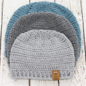 May include: Three crocheted beanies in shades of blue, grey, and light grey are stacked. The beanies have a ribbed texture and a small brown tag with white text. The hats are arranged on a light-coloured wooden surface.