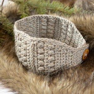 Puede incluir: Una diadema de ganchillo beige con un patrón texturizado de puntos verticales y un cierre de botón de madera marrón. El accesorio está elaborado con hilo de tono neutro y muestra un diseño de tejido consistente y repetitivo en toda la pieza.
