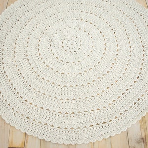 May include: A circular off-white crochet rug featuring a complex, repeating radial pattern. The design consists of concentric rings of intricate stitches, including solid bands and delicate, scalloped edges that form a decorative border.
