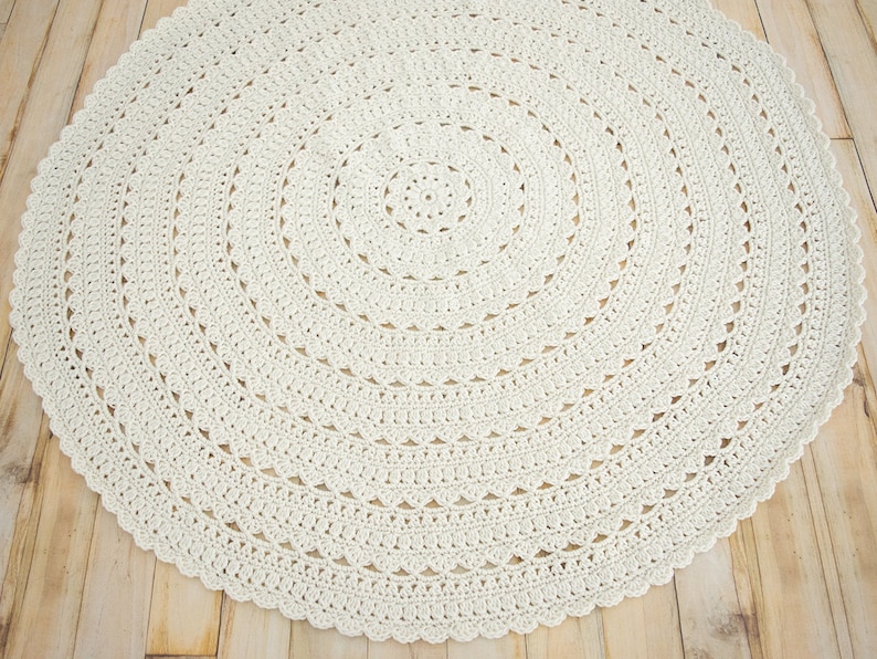 May include: A circular off-white crochet rug featuring a complex, repeating radial pattern. The design consists of concentric rings of intricate stitches, including solid bands and delicate, scalloped edges that form a decorative border.