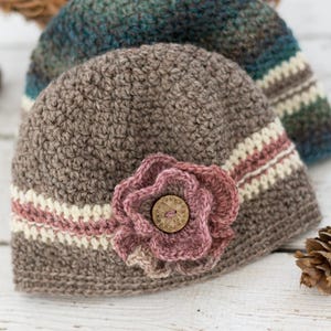 May include: A brown crocheted beanie with a pink crocheted flower and a button in the center. The beanie has a white and pink stripe around the bottom.