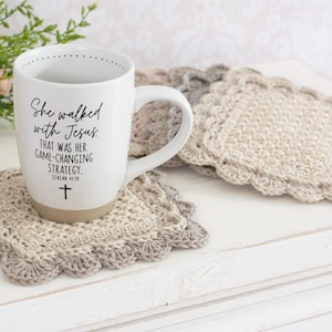 Könnte beinhalten: Weiße Keramiktasse mit dem Text "She walked with Jesus" und einem Kreuzsymbol. Die Tasse steht auf einem gehäkelten Untersetzer. Der Untersetzer ist beige und grau mit einem gewellten Rand. Ein zweiter Untersetzer ist sichtbar.