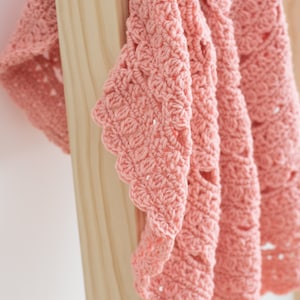 May include: A soft, salmon-pink crochet blanket featuring a repeating shell stitch pattern with scalloped edges. The textile displays a dense, textured construction with visible yarn loops and openwork details throughout the fabric design.