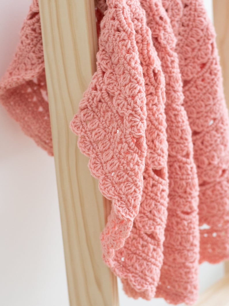 May include: A soft, salmon-pink crochet blanket featuring a repeating shell stitch pattern with scalloped edges. The textile displays a dense, textured construction with visible yarn loops and openwork details throughout the fabric design.