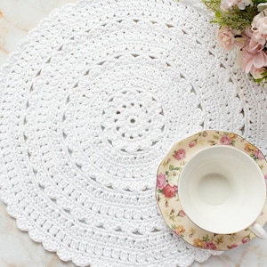 May include: A white, circular, crocheted doily with a scalloped edge. A floral teacup and saucer sit on the doily, next to a small bouquet of pink and white flowers. The doily is handmade and adds a touch of elegance.
