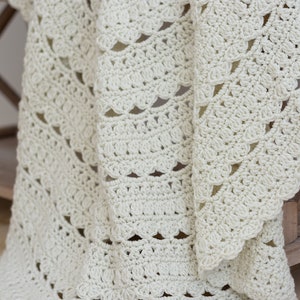 May include: An off-white crochet blanket featuring a repeating pattern of textured shell stitches and openwork rows. The fabric displays a consistent, intricate hand-stitched design with a scalloped edge, showcasing the soft yarn texture throughout.