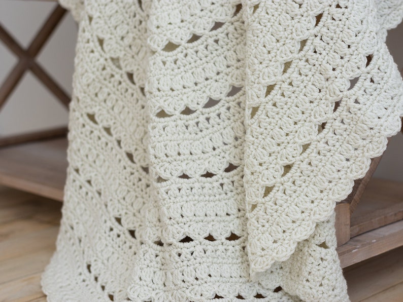 May include: An off-white crochet blanket featuring a repeating pattern of textured shell stitches and openwork rows. The fabric displays a consistent, intricate hand-stitched design with a scalloped edge, showcasing the soft yarn texture throughout.