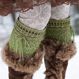 May include: Green crochet leg warmers with brown accents and faux fur trim. The leg warmers are paired with brown suede boots. The boots have metal ring details and are laced up the side. The background is a snowy outdoor scene.