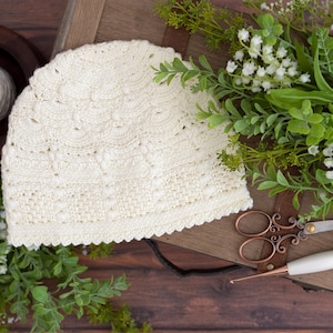 Puede incluir: Gorro de ganchillo color crema con borde festoneado, sobre una caja de madera. Una madeja de hilo está en un cuenco de madera. Vegetación, tijeras decorativas y un ganchillo completan la escena. El gorro está hecho a mano.