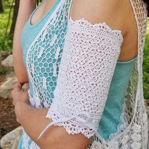 Può includere: Uno scaldabraccia in uncinetto bianco caratterizzato da un intricato motivo in pizzo con bordi smerlati sia in alto che in basso. Un sottile nastro bianco è annodato in un piccolo fiocco sul bordo inferiore della manica, indossata sopra un top azzurro.