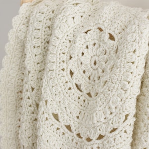 May include: An off-white crochet blanket featuring a circular mandala-inspired pattern with intricate stitching and a scalloped edge. The textile displays a repeating floral motif created with dense yarn loops, showcasing a textured surface.