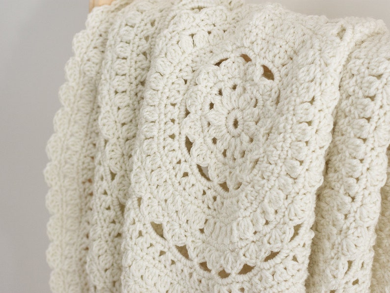 May include: An off-white crochet blanket featuring a circular mandala-inspired pattern with intricate stitching and a scalloped edge. The textile displays a repeating floral motif created with dense yarn loops, showcasing a textured surface.