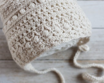 May include: A cream-colored crochet bonnet featuring a textured stitch pattern with small bobbles and an openwork design. The bonnet includes long, thin crocheted ties extending from the sides to secure the head covering in place.