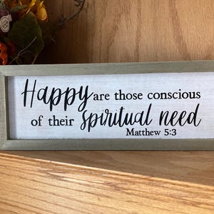 May include: A rectangular wooden sign with a light green frame. The sign has the words "Happy are those conscious of their spiritual need" in black script. The text "Matthew 5:3" is at the bottom.