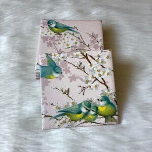 May include: Set of four pink coasters with a floral design featuring blue and yellow birds. The birds are perched on branches with white blossoms.
