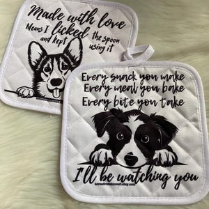 May include: A set of two white quilted pot holders with black dog illustrations and text. The first pot holder features a corgi with the text "Made with love. Means I licked the spoon and kept using it." The second pot holder features a border collie with the text "Every snack you make. Every meal you bake. Every bite you take. I'll be watching you."