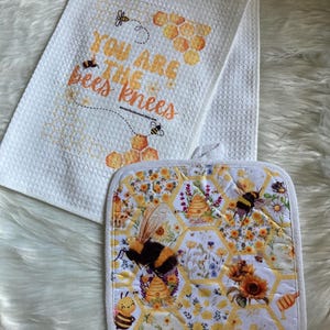 May include: A white kitchen towel and pot holder set with a bee theme. The towel has the text "YOU ARE THE bees knees" with bee and honeycomb graphics. The pot holder features bees, sunflowers, and honeycomb patterns.