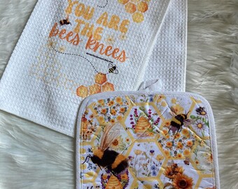 May include: A white kitchen towel and pot holder set with a bee theme. The towel has the text "YOU ARE THE bees knees" with bee and honeycomb graphics. The pot holder features bees, sunflowers, and honeycomb patterns.