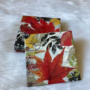 May include: A stack of four square coasters with a fall leaf design. The coasters feature a variety of red, yellow, and green leaves on a white background with black script.