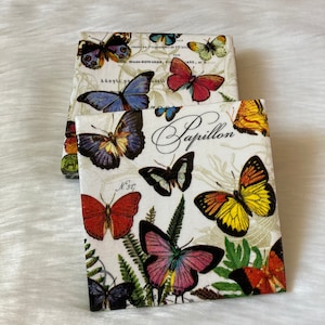 May include: A set of four white coasters with a butterfly design. The coasters feature colorful butterflies with black outlines and green leaves. The text "Papillon" is written in black cursive on the coasters.