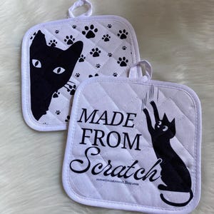 May include: Two white quilted pot holders with black cat designs. One pot holder has a cat peeking out from behind a paw print pattern and the other has a cat scratching at a paw print pattern. The text "MADE FROM Scratch" is printed on the second pot holder.