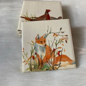 May include: Set of four ceramic coasters featuring a fox and a pheasant in a woodland setting. The coasters are white with a watercolor-style design.