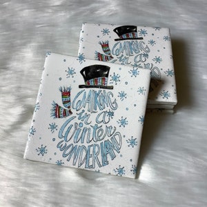 May include: Two white ceramic coasters with a snowman design and the text "Walking in a winter wonderland". The coasters have a white background with blue snowflakes.