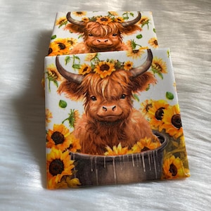 May include: Two square coasters with a white background and a brown and white Highland cow with a sunflower crown sitting in a pot of sunflowers.