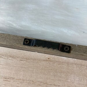 May include: A close-up of a wooden frame with a black metal hanging hook attached to the back. The hook has two screws and a serrated edge.
