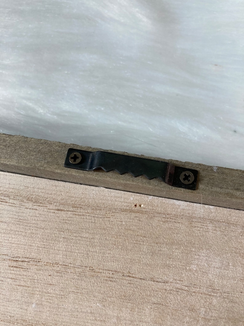 May include: A close-up of a wooden frame with a black metal hanging hook attached to the back. The hook has two screws and a serrated edge.