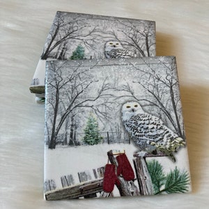 May include: Set of four ceramic coasters featuring a snowy winter scene with a white owl perched on a fence post. Red mittens hang from the fence.