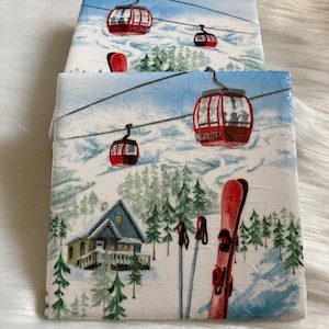 May include: Two ceramic coasters with a winter scene featuring red cable cars, a small cabin, and ski poles on a snowy mountainside.