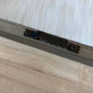 May include: A close-up of a wooden frame with a black metal hanging hook attached to the back. The hook has two screws and a serrated edge.