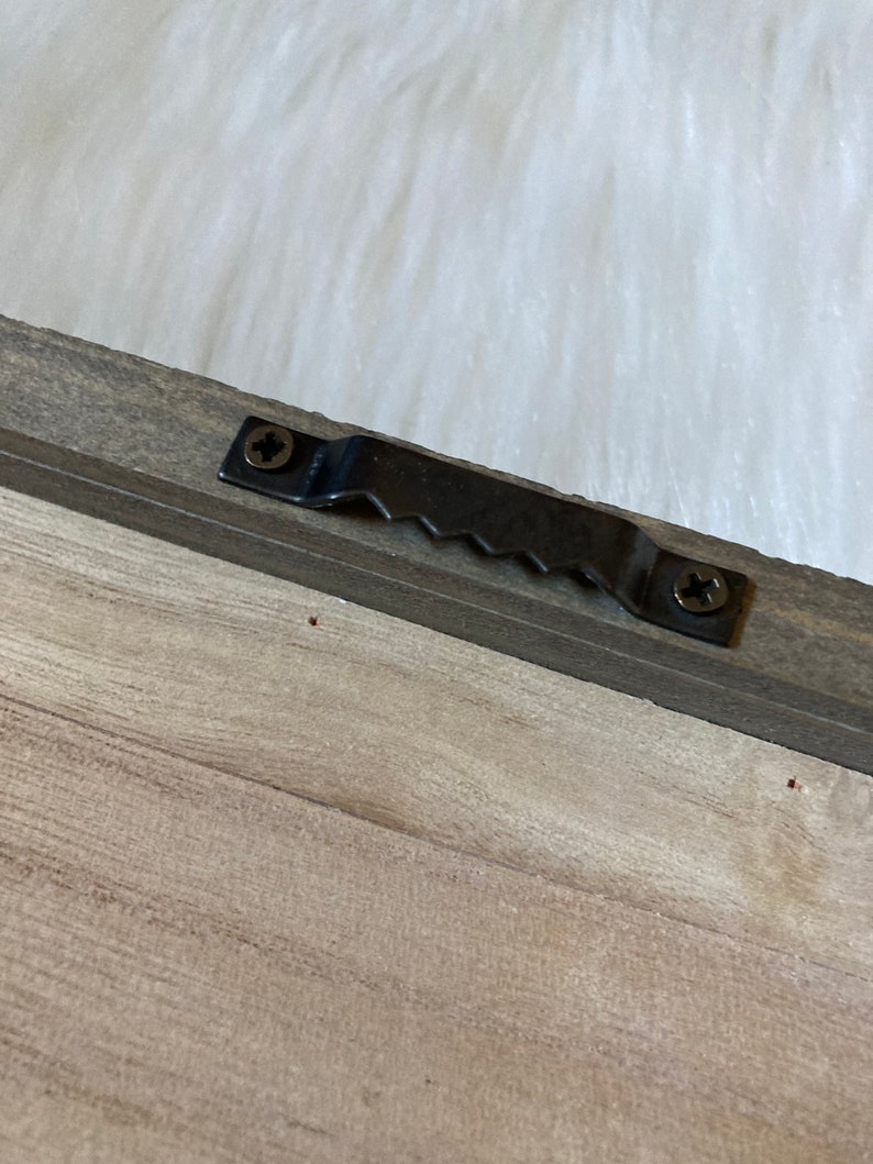 May include: A close-up of a wooden frame with a black metal hanging hook attached to the back. The hook has two screws and a serrated edge.